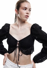 Blusa negra corta con mangas abullonadas, que presenta un patrón de ojal texturizado y un cierre delantero con nudo. Combinada con pantalones blancos.