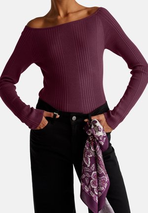 Personne portant un haut violet côtelé à manches longues et épaules dénudées, et un pantalon noir avec une écharpe violette à motifs nouée à la taille.