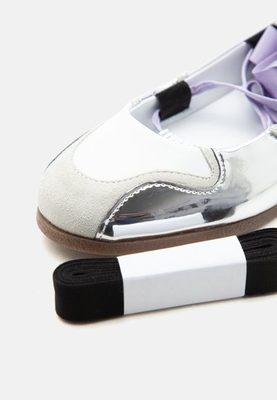 Gros plan d'une chaussure plate de ballet blanche et argentée avec un bout en daim et un lacet noir séparé enveloppé dans une bande de papier blanche.