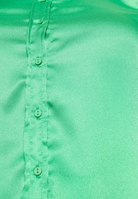 Blusa verde de cetim com uma pala de botões na frente, apresentando seis botões combinando. O tecido possui uma textura suave com um ligeiro brilho.