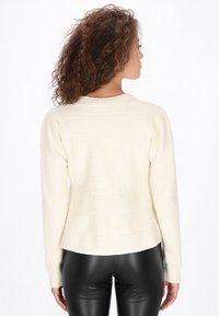 Weißer Strickpullover mit geripptem Kragen, langen Ärmeln und strukturiertem Körper. Kombiniert mit engen schwarzen Lederhosen.