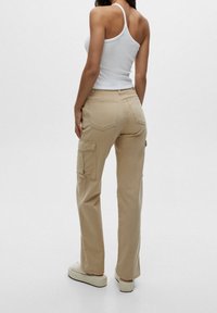 Beige cargo kalhoty vyrobené z bavlněné směsi; mají boční kapsy, rovný střih nohavic a střední výšku pasu. Nosené s bílým tílkem.