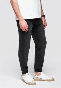 Mustat kapenevat farkut, joissa on joustava hihna, yhdistettyinä valkoisiin ja beigeihin tennareihin. Kangas on sileäpintaista ja minimalistista muotoilua.