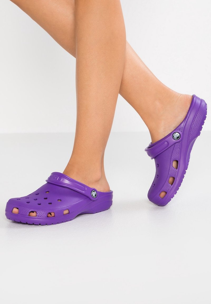 Jambes portant des sabots violets à enfiler avec des trous de ventilation, des semelles texturées et des sangles de talon réglables sur un fond uni.