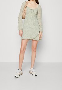 Ljusgrön klänning med blommönster, fyrkantig halsringning och långa genomskinliga puffärmar. Kombinerad med vita sneakers och en beige väska.