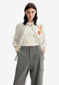 Camicia a righe bianca a maniche lunghe con ricami di fiori arancioni, abbinata a pantaloni cargo grigi. Top a costine di colore neutro sotto.