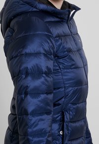 Person iført en marineblå, skinnende quiltet pufferjakke med vandret quiltning, lynlås, snap-knap lomme og fastgjort hætte.