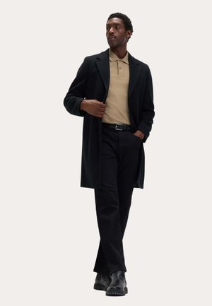 Mand iført en sort lang frakke, beige polo, sorte bukser, sort bælte og sorte støvler, stående med den ene hånd i lommen og den anden, der justerer frakken.