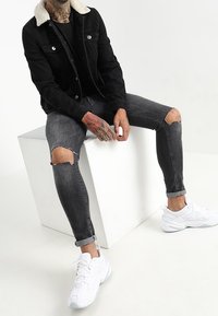 Černá džínová bunda s límcem z umělého ovčího rouna, doplněná o roztrhané šedé džíny a bílé sportovní tenisky. Sedí na bílém krychlovém bloku.