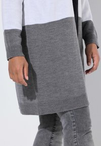 Person in einem langen grau-weißen Strickpullover und grauen Jeans, mit entspannten Händen an den Seiten vor einem schlichten Hintergrund.