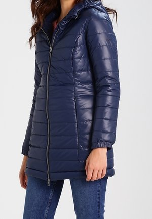 Femme portant une veste matelassée à capuche bleu marine avec fermeture éclair à l'avant et poignets élastiques, associée à un jean en denim bleu.