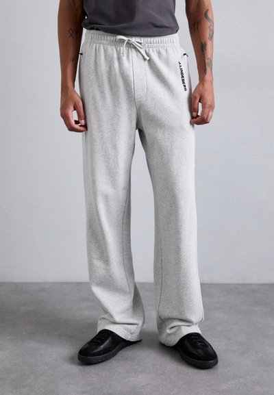 Homme portant un pantalon de survêtement gris clair avec un élastique à la taille et un cordon de serrage, des baskets noires, debout contre un fond gris clair uni.