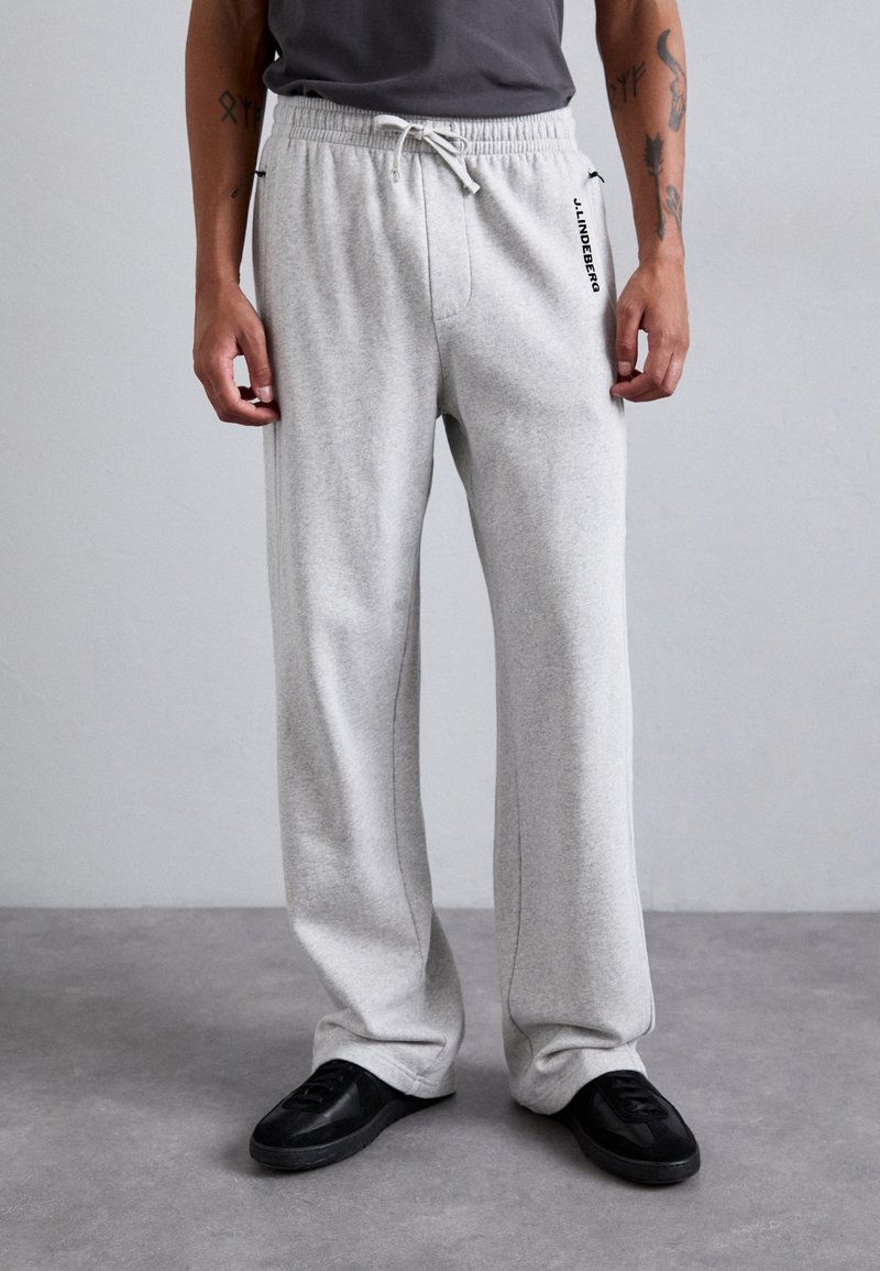 Man wearing light gray sweatpants with elastic waistband and drawstring, black sneakers, standing against plain light gray background.