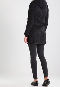 Persoon in een zwarte hoodie, zwarte skinny broek en zwarte glanzende loafers, staand met de rug naar de camera tegen een effen witte achtergrond.
