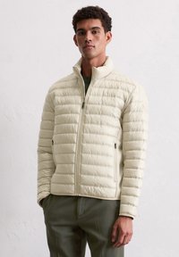 Kremowa pikowana kurtka puchowa z poziomym wzorem pikowania, wysokim kołnierzem, zamkiem błyskawicznym z przodu i bocznymi kieszeniami, wykonana z błyszczącego materiału.
