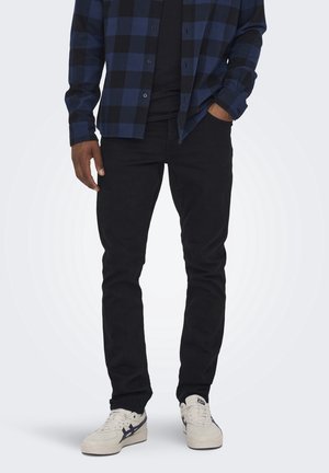 Homme portant un pantalon noir, des baskets blanches et une chemise à carreaux bleue et noire, une main dans la poche, sur un fond uni.