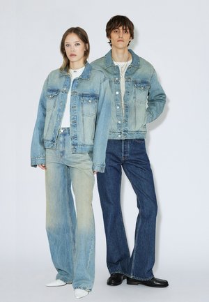 Kaksi nuorta aikuista seisoo vierekkäin, pukeutuneina vaaleansinisiin farkkutakkeihin, korkeavyötäröisiin farkkuihin, valkoisiin toppeihin sekä mustiin tai valkoisiin kenkiin.