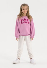 Rosa Sweatshirt mit dem Aufdruck "HELLO SUMMER", kombiniert mit weißen Jogginghosen mit Seitentaschen und weißen Sneakers. Glatte, weiche Texturen.