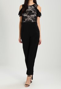 Czarna koronkowa bluzka z odkrytymi ramionami, wzory kwiatowe, połączona z czarnymi spodniami o wysokim stanie i straponami na wysokim obcasie.