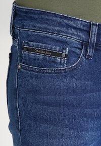 Close-up of blue denim Calvin Klein jeans pocket with visible stitching, belt loop, and small black label on coin pocket.