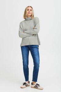 Pullover en tricot léger gris, coupe décontractée, associé à un jean slim bleu et des baskets beiges avec des accents noirs, se tenant contre un fond blanc.