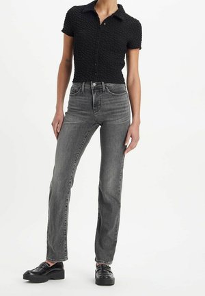 Kvinde iført en kortærmet sort tekstureret skjorte med knapper, højtsiddende falmede grå jeans og sorte platåsko på hvid baggrund.