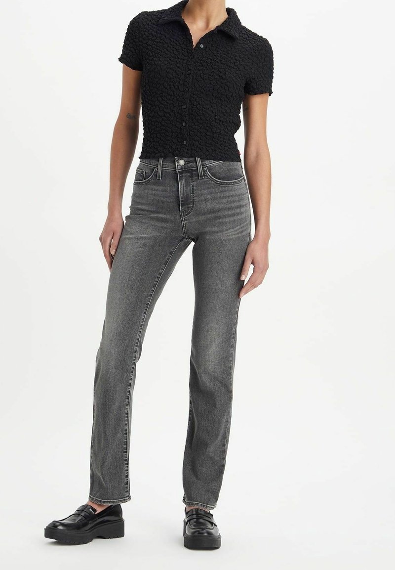 Femme portant une chemise noire texturée à manches courtes et boutonnée, un jean gris délavé taille haute et des mocassins à plateforme noirs sur fond blanc.