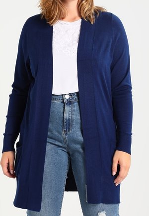 Femme portant un long cardigan bleu marine sur un chemisier blanc brodé et un jean bleu taille haute avec une petite déchirure au genou.