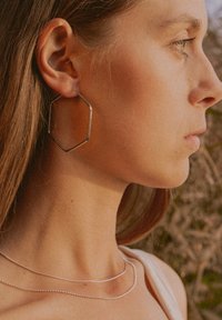 Silberne sechseckige Creolen und zwei zarte Halsketten – eine flache und eine mit Perlen – getragen von einer Person mit langen Haaren.