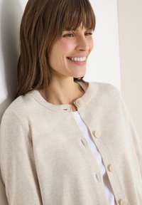 Femme aux cheveux bruns souriante, portant un cardigan beige boutonné sur un haut blanc, légèrement appuyée contre un mur blanc.