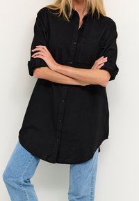 Chemise noire boutonnée en tissu léger, dotée d'une poche poitrine unique, manches retroussées et ourlet arrondi. Portée avec un jean bleu clair.