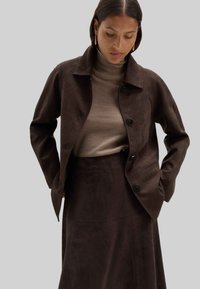 Kvinna iklädd en mörkbrun mockajacka och kjol med en beige polotröja, tittar ner med händerna i jackfickorna mot en grå bakgrund.
