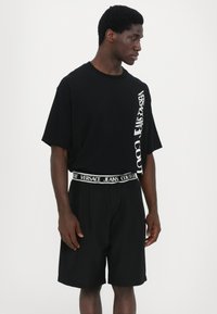 Svart t-skjorte med korte ermer som har en hvit "Versace Jeans Couture" logo på siden, kombinert med matchende svarte shorts og en merket midjebånd.