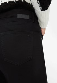 Zwarte spijkerbroek van denim met een gladde textuur, voorzien van een achterzak en een leren label. Het ontwerp is minimalistisch met subtiele stikseldetails.