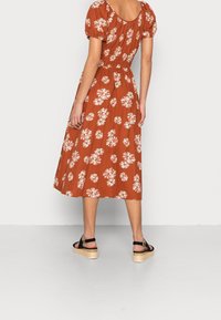 Robe midi couleur rouille avec des motifs floraux blancs, manches courtes bouffantes, taille élastique et jupe évasée. Associée à des sandales noires à plateforme.