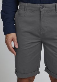 Gray cotton shorts with rolled cuffs, featuring side pockets and a button closure, worn with a navy shirt, hand resting on the thigh.