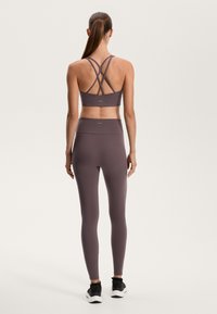 Ljubičasti sportski grudnjak s dizajnom u obliku križa na leđima, uparen s matching tajicama, oboje napravljeno od glatkog, rastezljivog materijala. Crne atletske tenisice.