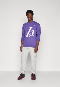 Sweatshirt roxo com gráfico branco "LA", combinado com calças de fato cinza. O modelo usa sapatilhas pretas e vermelhas, exibindo um look casual.