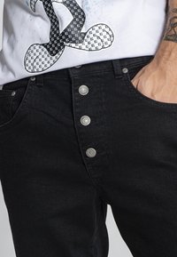 Jeans de mezclilla negra con cierre de cinco botones, herrajes metálicos y un corte clásico. Textura suave con un ligero desgaste y costuras en contraste.