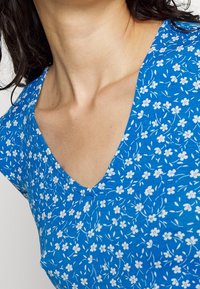 Femme portant un t-shirt bleu en V avec un motif floral blanc, montrant son cou et le haut de sa poitrine, avec des cheveux bouclés foncés visibles.