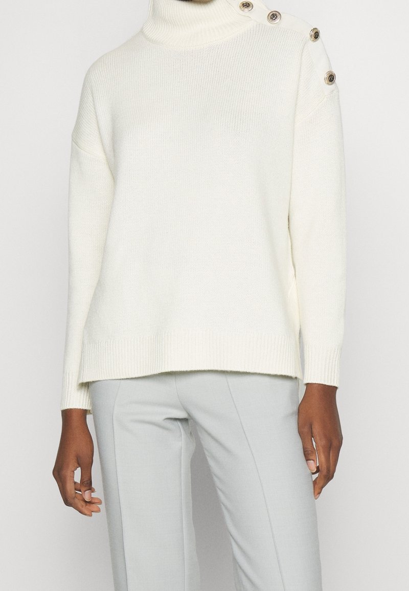 Kvinna iklädd en vit stickad tröja med tre dekorativa knappar på axeln och ljusgrå skräddarsydda byxor.