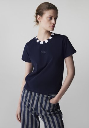 Mujer con una blusa de manga corta azul marino con cuello a rayas blancas y azules marinas, y pantalones a rayas verticales azules y grises, con la mano en el bolsillo.