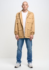 Chaqueta utility beige con múltiples bolsillos, camiseta blanca debajo, jeans azules y zapatillas deportivas grises. Modelo de pie contra un fondo claro.