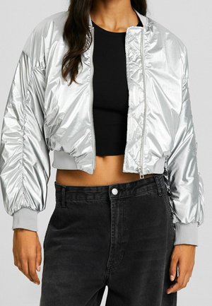 Femme portant une veste bomber argentée brillante courte sur un crop top noir et un jean noir taille haute, debout devant un fond uni.