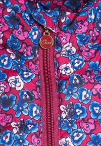 Närbild av en rosa jacka med ett blått och vitt blommönster samt en röd dragkedjeanordning med texten "Emoi" ingraverad.