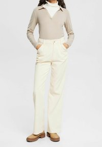 Maglietta beige a maniche lunghe con scollatura a V, abbinata a pantaloni in corda crema a vita alta con linee verticali e tasche laterali. Zoccoli marroni.