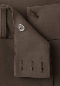 Hnědý manžetový okraj bundy s průhledným knoflíkem a třemi svislými knoflíkovými dírkami na texturované látce, detail.