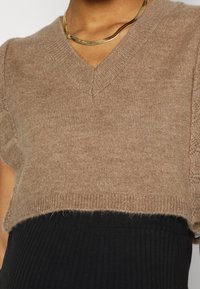 Brązowy, krótki sweter dzianinowy z dekoltem w serek, krótkimi rękawami i prążkowanymi krawędziami, noszony na czarnej sukience prążkowanej.