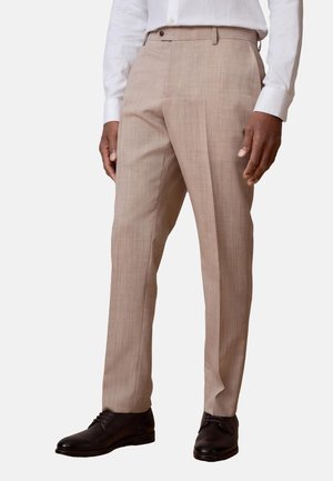Hombre con pantalones de vestir beige, camisa blanca y zapatos de vestir negros, de pie contra un fondo claro y liso.