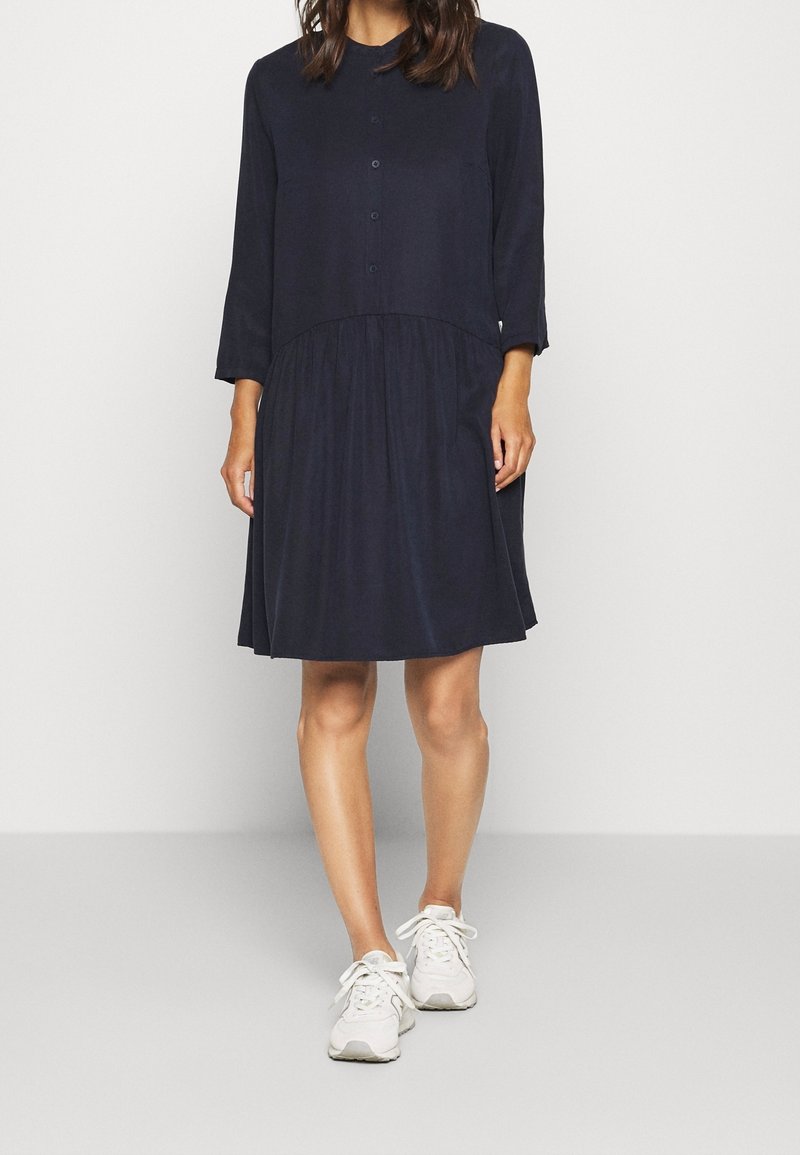 Donna che indossa un vestito blu scuro fino al ginocchio con maniche a tre quarti e bottoni nella parte superiore anteriore, abbinato a sneakers bianche, in piedi su uno sfondo semplice.